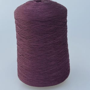 May include: A large, cylindrical spool of dark plum-colored yarn. The thread is tightly wound in a crisscross pattern around the central core, creating a uniform, textured surface across the entire height of the spool.