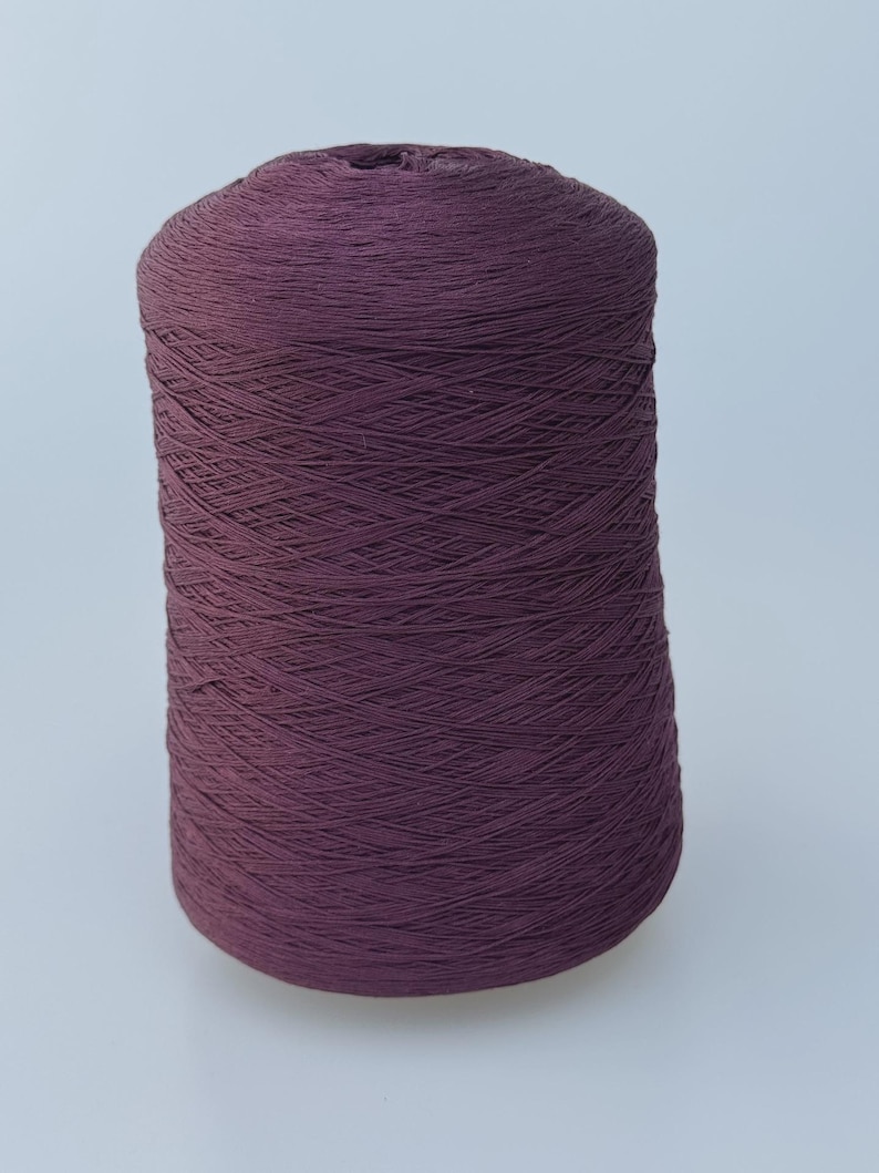 May include: A large, cylindrical spool of dark plum-colored yarn. The thread is tightly wound in a crisscross pattern around the central core, creating a uniform, textured surface across the entire height of the spool.