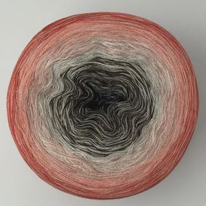 May include: A circular cake of yarn featuring a gradient color transition. The center is a deep black that shifts into shades of light gray, then transitions into a vibrant coral pink at the outer edges. The yarn is wound in a tight, concentric spiral.