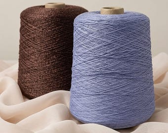 May include: Two large conical spools of yarn, one in a deep chocolate brown and the other in a soft lavender blue, each featuring a central cardboard core, standing upright and neatly wound with a consistent, fine-textured thread pattern.