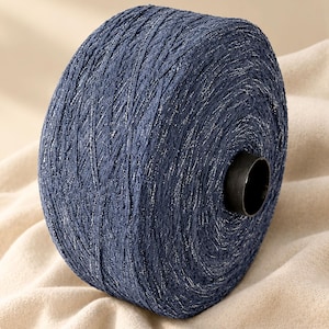 May include: A large spool of dark navy blue yarn featuring a metallic silver thread woven throughout the fibers. The yarn is wound tightly onto a black cylindrical core, creating a textured, shimmering appearance across the entire surface.