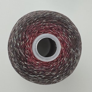 May include: A circular spool of yarn featuring a variegated color palette of deep burgundy, dark charcoal, and black, interwoven with thin, shimmering silver metallic threads throughout the fiber, centered around a clear plastic cylindrical core.