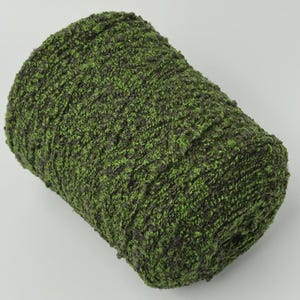 May include: A cylindrical skein of yarn featuring a textured, boucle-style construction. The yarn displays a variegated color palette, blending deep forest green and vibrant lime green fibers throughout the entire length of the wound material.