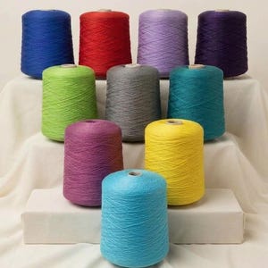May include: Ten cylindrical spools of yarn arranged in a tiered display. The colors include royal blue, red, light purple, dark purple, lime green, gray, teal, magenta, yellow, and light blue. Each spool features a visible cross-hatch thread pattern.