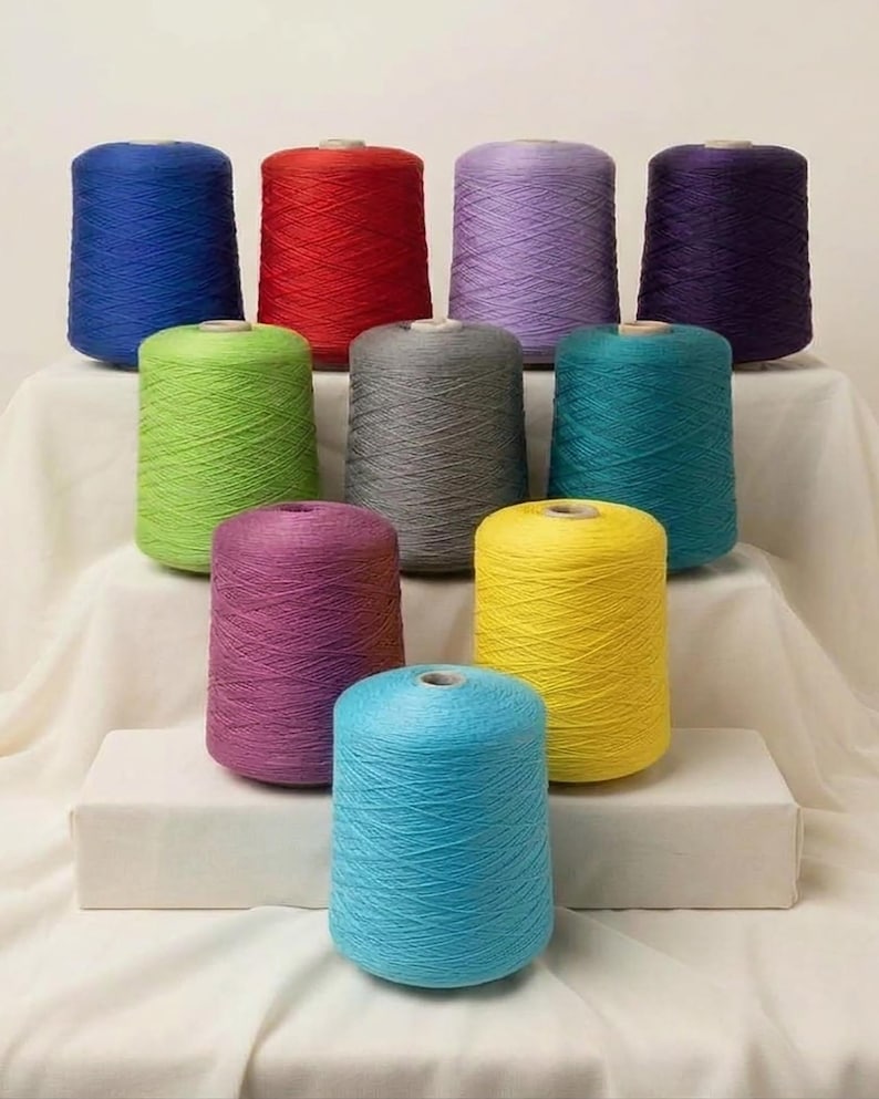 May include: Ten cylindrical spools of yarn arranged in a tiered display. The colors include royal blue, red, light purple, dark purple, lime green, gray, teal, magenta, yellow, and light blue. Each spool features a visible cross-hatch thread pattern.