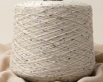 May include: A large conical spool of off-white yarn featuring a speckled pattern with small flecks of blue, brown, and black throughout the thread. The yarn is tightly wound around a central core, with a green circular top visible at the center.
