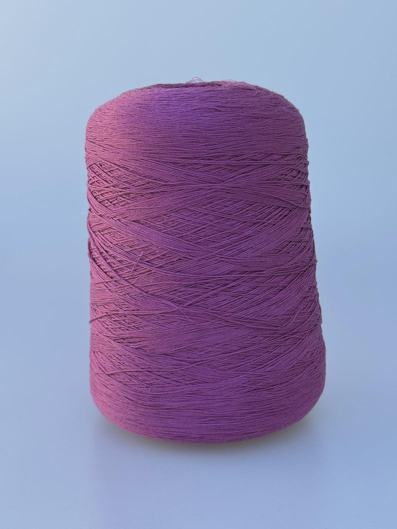 May include: A large, cylindrical spool of deep purple yarn. The thread is tightly wound in a crisscross pattern around the central core, creating a textured surface. The spool stands upright, displaying a consistent, rich violet hue throughout.