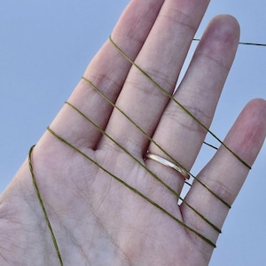 May include: A thin, olive green thread is wrapped multiple times around the fingers of an open hand. The thread is smooth and uniform in color, creating a series of parallel lines across the skin. The texture of the thread appears fine.