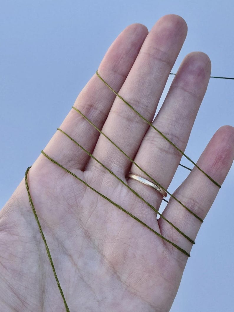 May include: A thin, olive green thread is wrapped multiple times around the fingers of an open hand. The thread is smooth and uniform in color, creating a series of parallel lines across the skin. The texture of the thread appears fine.