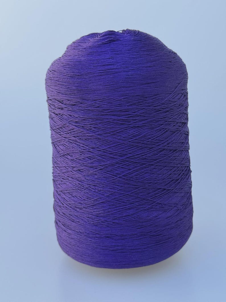 May include: A cylindrical spool of deep purple yarn wound tightly in a cross-hatch pattern. The thread is thin and uniform, creating a smooth, dense texture across the entire surface of the spool, which stands upright against a plain background.
