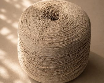May include: A cylindrical spool of light beige yarn featuring a tightly wound, textured thread construction. The yarn displays a consistent neutral tan color throughout the entire spool, with a visible central opening at the top of the structure.