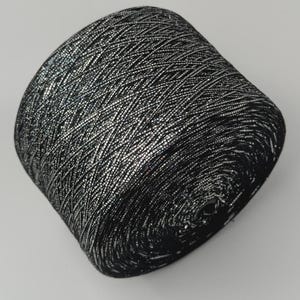 May include: A cylindrical spool of yarn featuring a metallic silver and black color palette. The thread is wound in a tight, crisscross pattern, creating a textured, shimmering surface that reflects light across the entire surface of the spool.