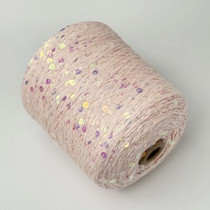 May include: A cylindrical spool of pale pink yarn featuring scattered iridescent sequins that reflect light in various colors. The yarn is wound tightly around a central cardboard core, creating a textured surface with a subtle shimmer throughout.
