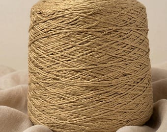 May include: A large, cylindrical spool of golden-tan yarn featuring a tightly wound, textured fiber construction. The yarn exhibits a subtle metallic sheen and a consistent, braided appearance throughout the entire spool, creating a uniform surface.