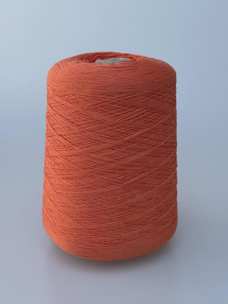 May include: A large, cylindrical spool of vibrant terracotta-colored yarn. The thread is tightly wound in a cross-hatch pattern around a central core, creating a uniform, textured surface that tapers slightly toward the top of the spool.