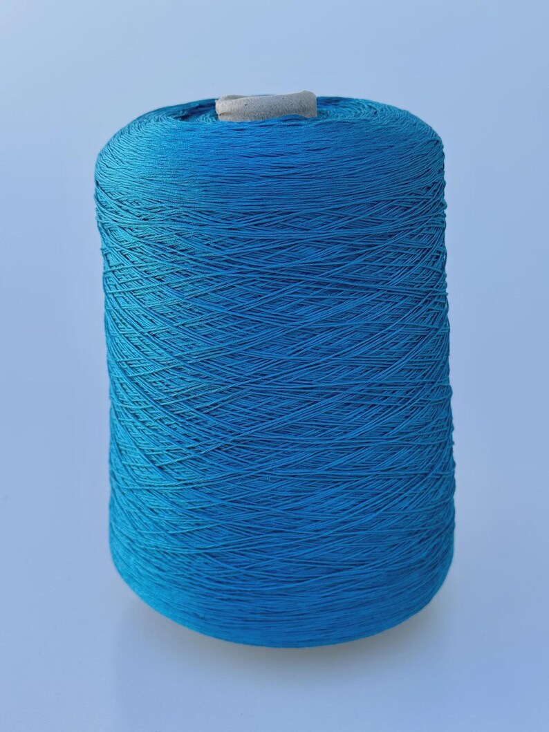 May include: A large cylindrical spool of vibrant turquoise blue yarn. The thread is tightly wound in a crisscross pattern around the central core, which is visible at the top. The spool has a smooth, uniform shape and a consistent bright blue color.