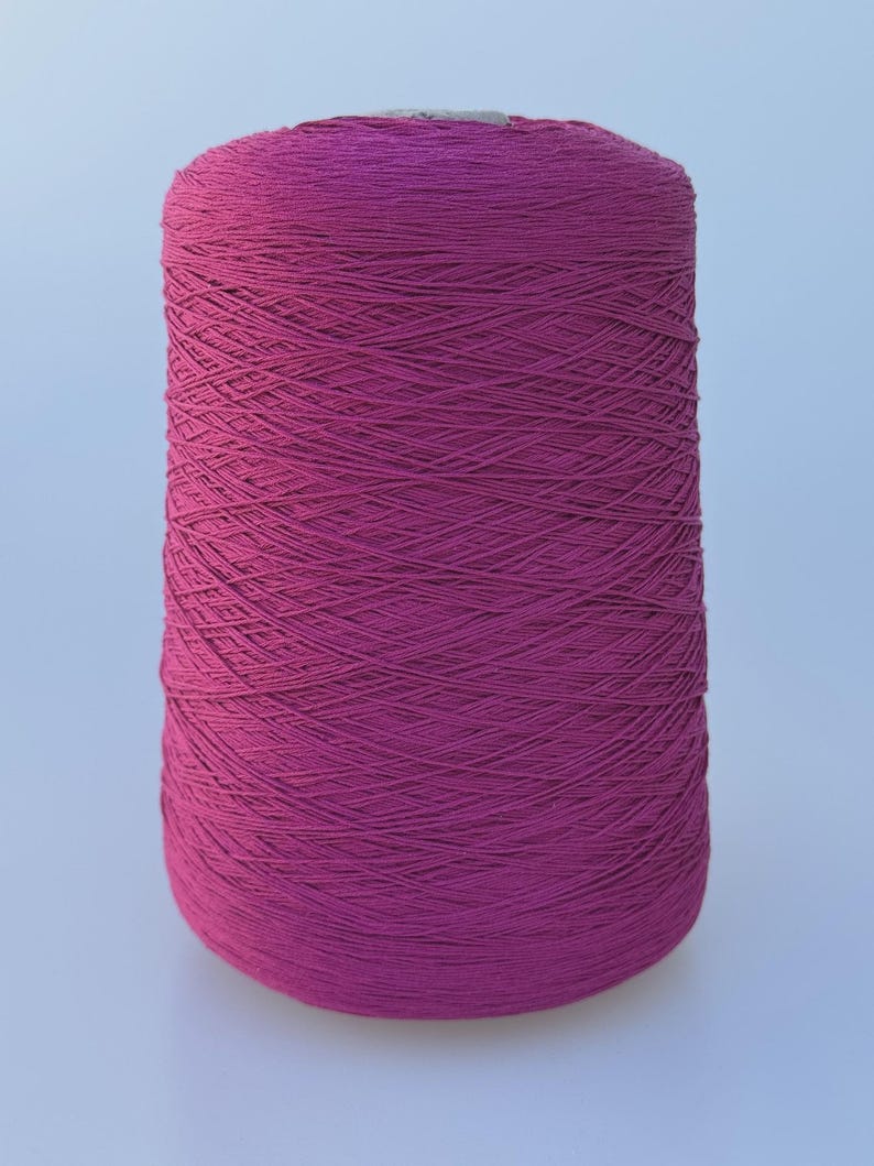 May include: A large cylindrical spool of vibrant magenta yarn. The thread is tightly wound in a crisscross pattern around the central core, creating a textured surface with a consistent deep pink color throughout the entire spool.