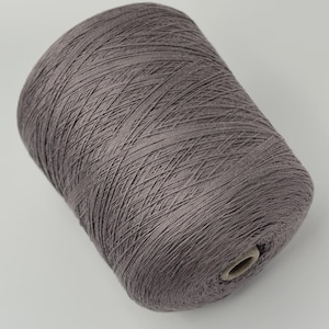 May include: A large, cylindrical spool of dusty mauve-colored yarn. The thread is tightly wound in a cross-hatch pattern around a central cardboard core, creating a uniform, textured surface across the entire exterior of the yarn cone.