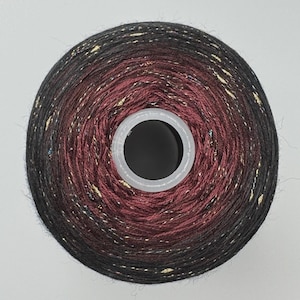 May include: A circular spool of yarn featuring a gradient color transition from deep black on the outer edges to a rich burgundy red in the center, interspersed with fine metallic gold and iridescent blue flecks throughout the fiber texture.