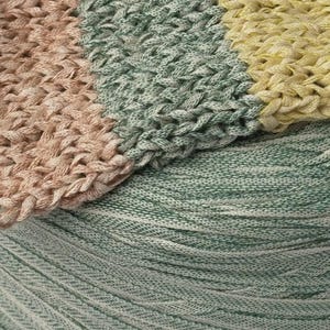 May include: A close-up view of a knitted textile featuring horizontal stripes in muted peach, sage green, and pale yellow. Below the knitted section, a large, tightly wound ball of sage green yarn shows a smooth, continuous fiber texture.
