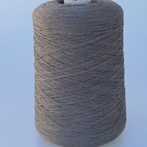 May include: A large cylindrical spool of taupe-colored yarn. The thread is tightly wound in a crisscross pattern around a central cardboard core, creating a uniform, textured surface across the entire exterior of the spool.