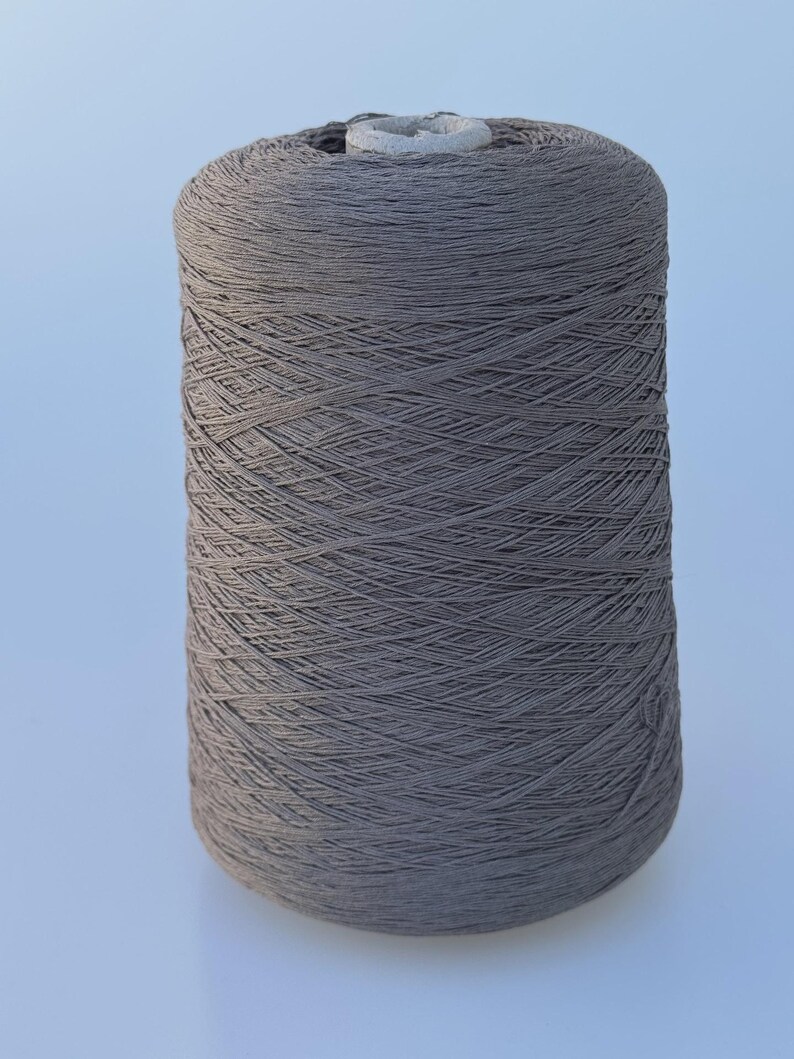 May include: A large cylindrical spool of taupe-colored yarn. The thread is tightly wound in a crisscross pattern around a central cardboard core, creating a uniform, textured surface across the entire exterior of the spool.