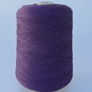 May include: A large cylindrical spool of deep purple thread wound tightly in a crisscross pattern. The spool features a light gray plastic core visible at the top center, providing a sturdy base for the densely packed, vibrant violet textile fiber.