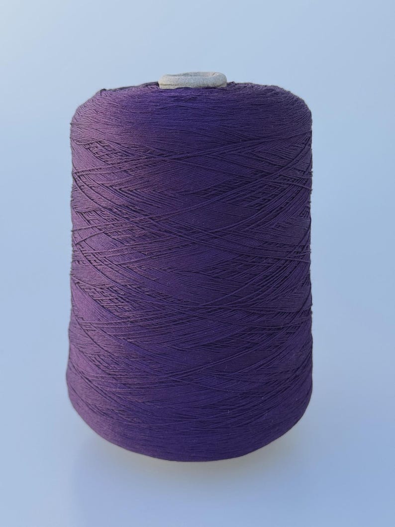 May include: A large cylindrical spool of deep purple thread wound tightly in a crisscross pattern. The spool features a light gray plastic core visible at the top center, providing a sturdy base for the densely packed, vibrant violet textile fiber.