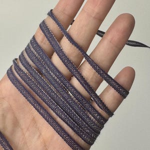 May include: A narrow, dark purple ribbon with a textured, woven appearance and a subtle metallic silver shimmer along the edges. The ribbon is wrapped multiple times in a continuous loop, showcasing its consistent width and detailed fabric weave.