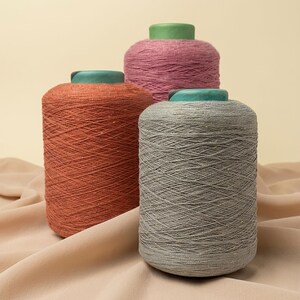 May include: Three cylindrical spools of yarn in shades of rust orange, dusty pink, and heather gray. Each spool features a solid green plastic core at the top, with the yarn wound tightly around the center in a consistent, textured pattern.