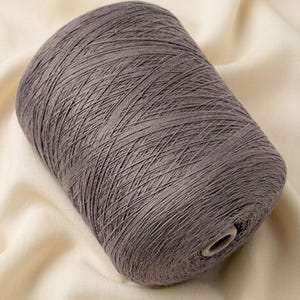 May include: A large, cylindrical spool of taupe-colored yarn. The yarn is tightly wound in a cross-hatch pattern around a central cardboard core, displaying a consistent, fine-textured thread throughout the entire surface of the spool.