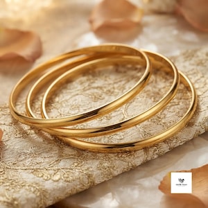 May include: Three polished gold bangles with a smooth, rounded surface. The bracelets are circular and feature a reflective, metallic finish. Each bangle is a simple, continuous band without any additional embellishments or textures.