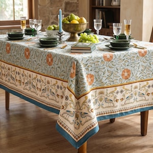 May include: A rectangular tablecloth featuring a light blue background with orange floral patterns and a cream-colored border decorated with blue and gold bird motifs. The table is set with green bowls, clear glassware, a fruit bowl, and a blue candle.