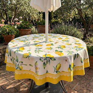 Könnte beinhalten: Eine runde Tischdecke mit einem Muster aus gelben Zitronen, grünen Blättern und weißen Blüten auf weißem Grund. Die Tischdecke hat einen breiten gelben Rand mit einer weißen, bogenförmigen Spitzenborte am Saum und ein Loch in der Mitte.