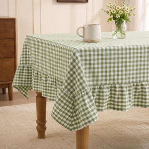May include: A rectangular tablecloth featuring a green and white gingham check pattern with a ruffled edge. A plain off-white ceramic mug sits on the surface, and a clear glass vase containing a bouquet of white daisies is placed nearby.