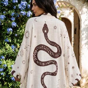 May include: A cream-colored kimono featuring a large brown snake graphic on the back, surrounded by various celestial symbols including suns, moons, and stars. The garment has wide sleeves, a textured fabric finish, and fringe trim along the hem.
