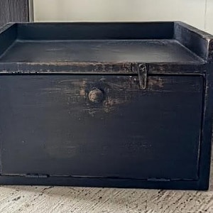 Puede incluir: Una pequeña caja de madera negra con un acabado envejecido. La caja tiene una puerta frontal con bisagras, un pomo y un pestillo. La parte superior de la caja tiene un pequeño borde alrededor de los bordes. La caja está sobre una superficie blanca.