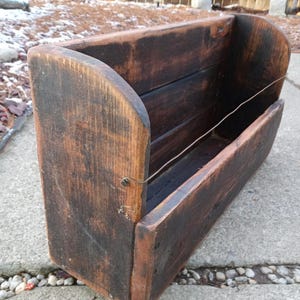 Może przedstawiać: Prostokątna drewniana donica o ciemnym, zwietrzałym wykończeniu. Donica ma zakrzywione boki i druciany detal, prezentując estetykę vintage. Widoczne usłojenie drewna dodaje naturalnego uroku.