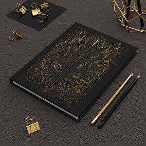 Puede incluir: Un cuaderno de tapa dura negra con una ilustración central de una hoja dorada que contiene dos ojos visibles entre el follaje. La cubierta está decorada con motas doradas. Dos lápices y varios clips de metal dorados se encuentran cerca.