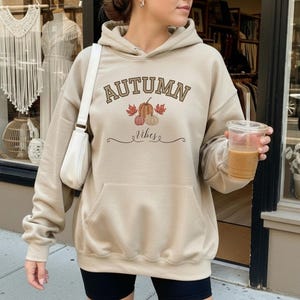 Könnte beinhalten: Ein beigefarbener Kapuzenpullover mit dem Text "AUTUMN" in brauner, gebogener Schrift über einer Grafik aus drei Kürbissen und Herbstblättern, dem Wort "Vibes" in Schreibschrift darunter und einer großen Kängurutasche vorne.