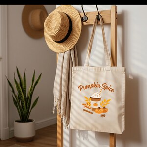 Könnte beinhalten: Eine beigefarbene Stofftasche mit einem grafischen Aufdruck eines Kaffeebechers mit Schlagsahne, einem Kürbis, Herbstblättern, einer Zimtstange und einem Sternanis, wobei der Text Pumpkin Spice in Orange über der Grafik gedruckt ist.