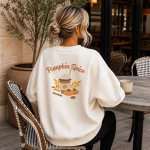 Könnte beinhalten: Ein cremefarbenes Sweatshirt mit Rundhalsausschnitt und einem Aufdruck auf der Rückseite mit dem Text Pumpkin Spice. Das Motiv zeigt einen Kaffeebecher mit Schlagsahne, Herbstblätter, zwei kleine Gebäckstücke, eine Zimtstange und einen Sternanis.