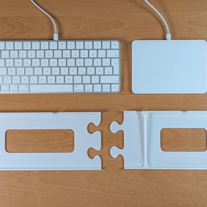 Puede incluir: Un teclado inalámbrico blanco y un trackpad blanco, cada uno con un cable de carga blanco, colocados sobre dos bandejas de plástico blanco con bordes de pieza de rompecabezas entrelazados y recortes rectangulares para los dispositivos.