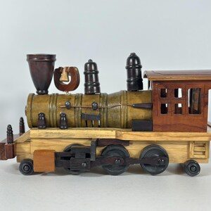 May include: A wooden toy train locomotive featuring a multi-toned finish with a light tan base, a mustard yellow cylindrical boiler, and a dark brown cab. It includes a dark funnel, a small bell, two black smokestacks, and black wheels.