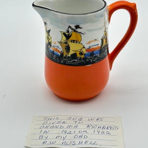 May include: A ceramic pitcher with a bright orange base and a handle, featuring a painted scene of yellow sailing ships on a blue sea around the upper rim. A handwritten note reads: THIS JUG WAS GIVEN TO GRANDMA RICHARDS IN 1921 OR 1922 BY MY DAD R.W. BUSHELL.