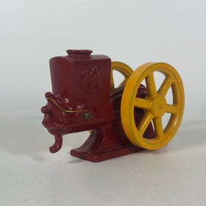 May include: A cast iron stationary engine model painted in deep red with a bright yellow flywheel. The engine features a molded eagle emblem on the side of the main body, a small metal crank handle, and a sturdy base, showcasing a mechanical design.