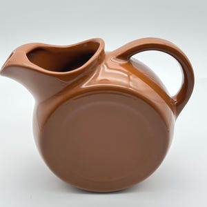 Peut inclure: Un pichet en céramique marron uni et brillant avec un corps rond et circulaire, un large bec verseur et une anse incurvée fixée sur le côté. La surface présente une finition lisse et réfléchissante sans aucun motif ou élément décoratif.