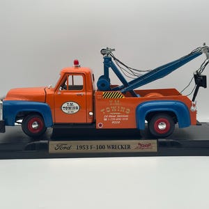 May include: A die-cast 1953 Ford F-100 wrecker model truck in orange and blue. The truck features Y.M. TOWING branding, a blue tow boom, a red roof light, and black wheels with red centres. It is mounted on a black display base with a nameplate.