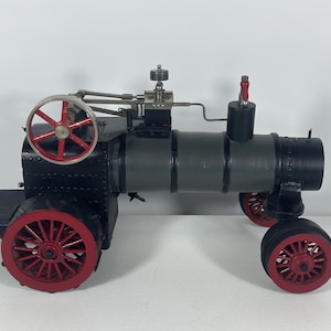 May include: A model steam tractor featuring a black and gray cylindrical boiler, a red spoked flywheel, and four red wheels. The engine assembly includes metallic components, a vertical chimney, and a red lever, all mounted on a black chassis.