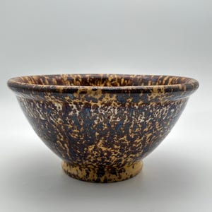 May include: A ceramic bowl featuring a speckled pattern in shades of dark brown, tan, and hints of blue. The bowl has a rounded rim, a deep basin, and a circular pedestal base, showcasing a mottled glaze finish across the entire exterior surface.